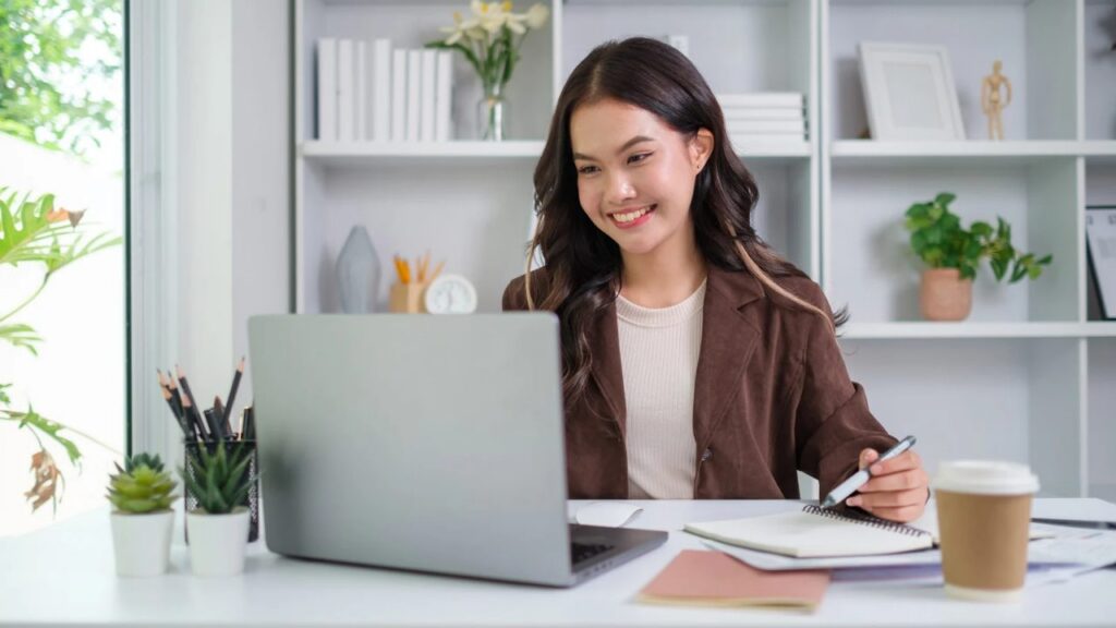 Smiling executive assistant in a brown blazer working at a laptop with a notebook