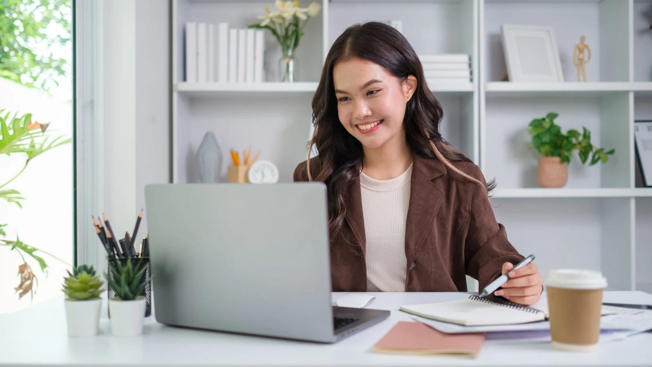 Smiling executive assistant in a brown blazer working at a laptop with a notebook